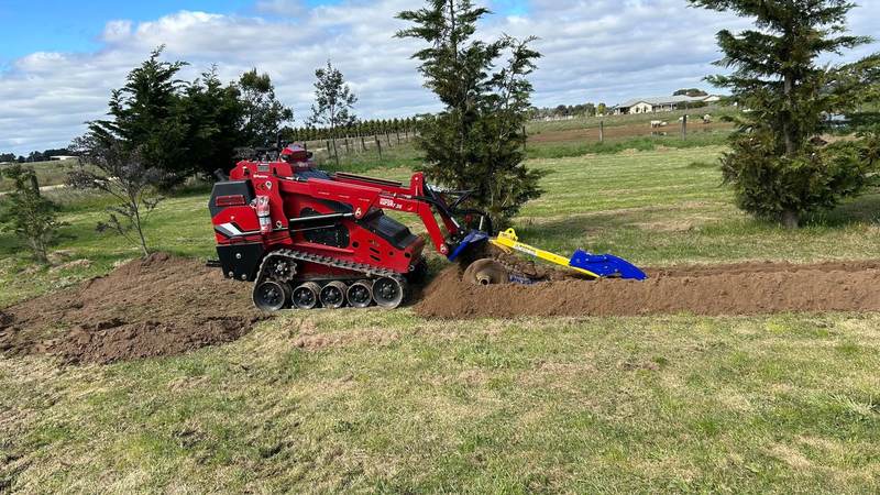 Red Darlington tracked mini loader trenching across a green lawn with trees